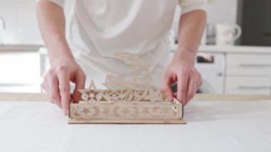 Genç, tanınmayan bir adam, masada duruyor, yan parçaları iki eliyle dipteki deliklere yerleştiriyor, oyulmuş ahşap bir tepsi birleştiriyor. Yakından, alttan yavaş çekimde. Konsept: tepsi montajı, Ramazan Kareem, Bayram Mübarek.