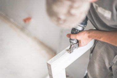 Olgun, tanımlanamayan beyaz şapkalı bir işçi, matkap kullanarak beyaz ahşap bir pencere çerçevesinden eski donanımı çıkarır. Yakın çekim, yukarıdan aşağıya manzara. Konsept: pencere iskeleti onarımı, teknoloji kullanımı.