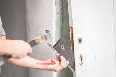 Olgun, beyaz bir işçi, elinde tornavida ve levye tutarken, eski ahşap bir kapıdan, yakın plan, yan görüş gibi mors kilidini kaldırıyor. Konsept: Tamir işi, kapı kilidi değiştirme.