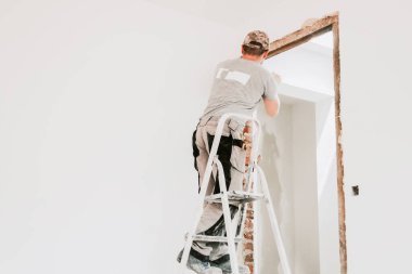 Üniformalı ve şapkalı genç bir beyaz adam, beyaz duvarlı bir odada sırtüstü duran, seçici odaklı, yakın çekimli bir odada, sırtından levye kullanarak eski macunu kapı aralığından temizliyor. İnşaat işi kavramı.