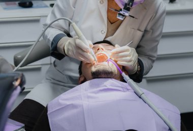 Genç beyaz bir adam, dişçi yüksek hızlı bir el yazısı, sonda ve tükürük püskürtücüsüyle tedavi prosedürünü uygularken gözleri kapalı bir şekilde diş sandalyesine uzanır. Yan görüş, yakın çekim. Konsept: Sözlü tedavi, dişçiler günü.