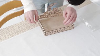 Genç bir beyaz adam yan parçalarını deliklere yerleştirir, hilal şeklinde oyulmuş ahşap bir tepsi, bir yıldız ve açık bir kitabı bir standın üzerine koyar. Yakın çekim, yavaş çekimde yan görüş. Konsept: tepsi montajı, Ramazan Kareem, Bayram Mübarek.