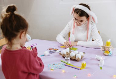 Tavşan bantlı iki beyaz kız kardeşin portresi, Paskalya yumurtalarını birlikte hazırlıyorlar, üzerine yapay elmaslar yapıştırıyorlar ve keçeli kalemle çiziyorlar, bir bahar günü mutfakta masada oturuyorlar, yan manzara yakın plan. Kavram