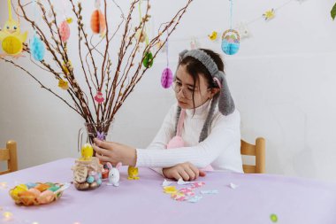 Tavşan bantlı güzel beyaz bir kızın portresi, masada oturmuş cam şeffaf sürahiye keçeli leylak etiketleri yapıştırıyor söğüt dalı ve asılı oyuncaklarla, bahar günü, yan tarafta Paskalya buketi yaratıyor.