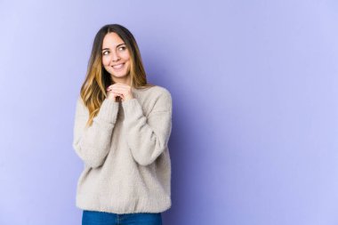 Mor arka planda izole edilmiş genç beyaz kadın ellerini çenesinin altında tutuyor, mutlu bir şekilde yana bakıyor..