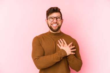 Pembe arka planda izole olmuş genç beyaz adam gülüyor, elini yüreğine koyuyor, mutluluk kavramını koruyor..