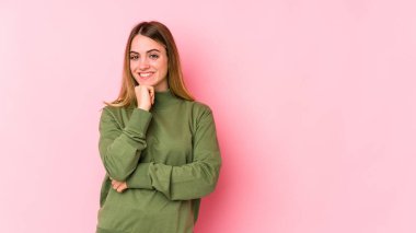 Pembe arka planda izole edilmiş genç beyaz kadın gülümsüyor ve kendinden emin, elleriyle çenesine dokunuyor..