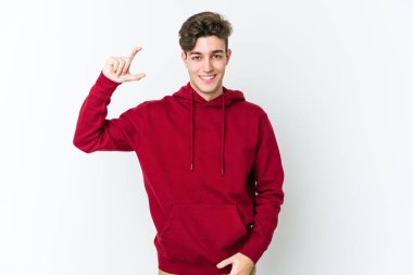 Beyaz arka planda izole edilmiş genç beyaz bir adam işaret parmaklarıyla küçük bir şey tutuyor, gülümsüyor ve kendinden emin..