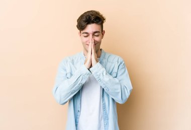 Beyaz tenli genç, bej arka planda izole edilmiş, el ele tutuşmuş, ağzı yakınında dua eden, kendinden emin..