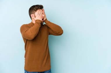 Mavi arka planda izole edilmiş genç beyaz adam elleriyle gözlerini kapatmaktan korkuyor..