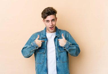 Beyaz tenli genç adam bej arka planda izole edildi. Parmakla göstererek şaşırdı, gülümsedi..