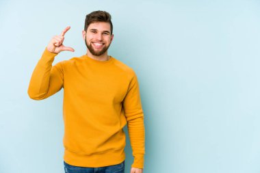 Kafkasyalı genç adam mavi arka planda izole edilmiş. Elinde işaret parmaklarıyla küçük bir şey tutuyor, gülümsüyor ve kendinden emin..