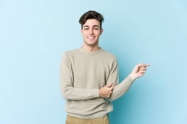 Mavi arka planda izole edilmiş genç beyaz adam işaret parmağıyla işaret ederek gülümsüyor..