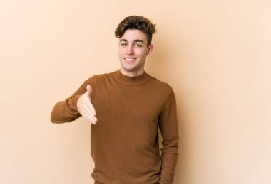 Beyazlı genç bir adam bej arka planda izole edilmiş. Kameraya doğru elini uzatarak selam veriyor..
