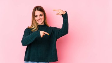 Pembe arka planda izole edilmiş genç beyaz bir kadın işaret parmaklarıyla küçük bir şey tutuyor, gülümsüyor ve kendinden emin..