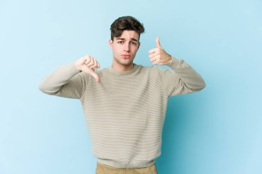 Kafkasyalı genç adam mavi arka planda izole edilmiş, baş parmağını kaldırıyor, zor seçim konsepti.