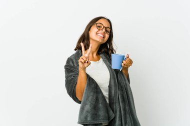 Beyaz arka planda battaniyeli genç Latin kadın zafer işareti gösteriyor ve gülümsüyor..