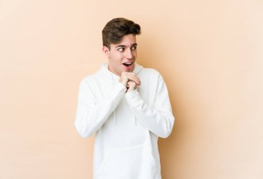 Beyaz tenli genç adam bej arka planda izole edilmiş şans için dua ediyor, şaşırmış ve ağzını açıp önüne bakıyor..