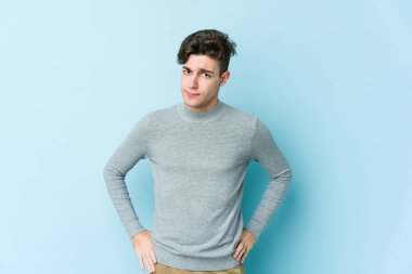 Kafkasyalı genç adam mavi arka planda izole, kafası karışmış, şüpheli ve güvensiz hissediyor..