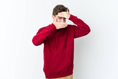 Beyaz arka planda izole edilmiş genç beyaz bir adam kameraya parmaklarıyla göz kırpıyor, yüzünü kapatmaktan utanıyor..