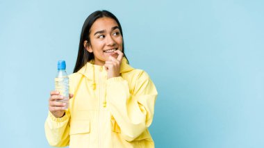 Genç Asyalı bir kadın mavi arka planda izole edilmiş bir şişe su tutuyor rahat bir şekilde fotokopi odasına bakan bir şeyi düşünüyor..