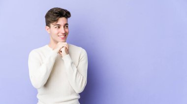 Mor arka planda izole edilmiş genç beyaz adam ellerini çenesinin altında tutuyor, mutlu bir şekilde yana bakıyor..