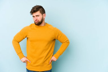 Kafkasyalı genç adam mavi arka planda izole, kafası karışmış, şüpheli ve güvensiz hissediyor..