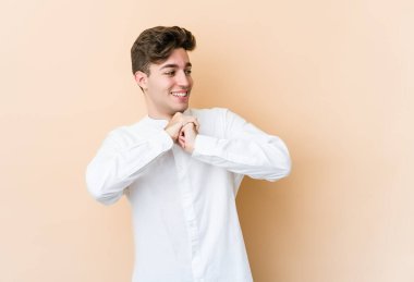 Beyaz tenli genç adam bej arka planda izole edilmiş ellerini çenesinin altında tutuyor, mutlu bir şekilde yana bakıyor..