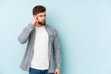 Mavi arka planda izole edilmiş genç beyaz bir adam şakağı parmağıyla işaret ediyor, düşünüyor ve bir göreve odaklanıyor..