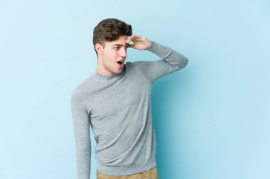 Kafkasyalı genç adam mavi arka planda izole edilmiş, uzaklara bakıyor ve elini alnına koyuyor..