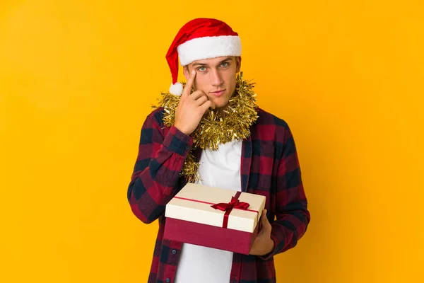 若いですCaucasian男とともにクリスマス帽子を保持していますプレゼント孤立した上に黄色の背景に聞こうとしているゴシップ — ストック写真