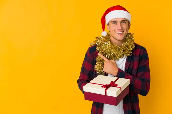 若いです白人男性とクリスマスの帽子を持つ存在を保持黄色の背景笑顔と脇を指して 空白のスペースで何かを示す — ストック写真