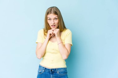 Mavi arka planda izole edilmiş genç sarışın kadın şans için dua ediyor, şaşırmış ve ağzı açık bir şekilde önüne bakıyor..