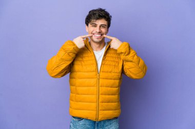 Genç havalı adam gülümsüyor, parmaklarıyla ağzını işaret ediyor..