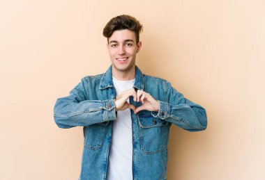 Beyaz tenli genç adam bej arka planda izole edilmiş gülümsüyor ve elleriyle kalp şeklini gösteriyor..