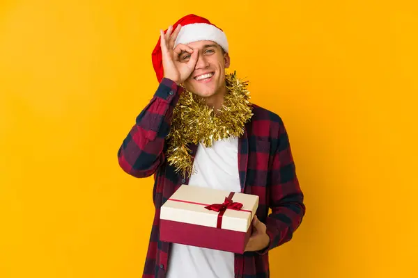 年轻的白种人头戴圣诞礼帽 拿着黄色背景的礼物 兴奋地保持着对着眼睛的正常姿势 — 图库照片