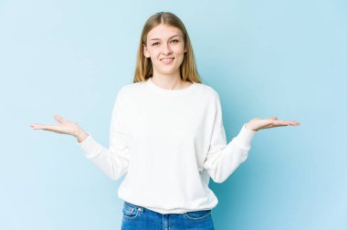 Mavi arka planda soyutlanmış genç sarışın kadın kollarıyla ölçeği yapıyor, mutlu ve kendinden emin hissediyor..