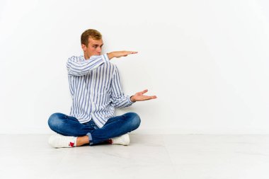 Yerde oturan genç beyaz adam şok oldu ve hayrete düştü. Ellerinin arasında bir kopya alanı tutuyor..