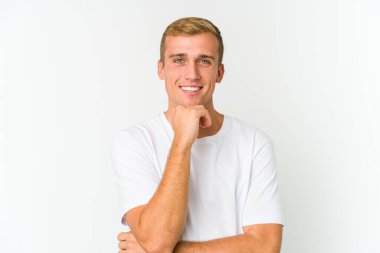 Genç beyaz yakışıklı adam gülümsüyor mutlu ve kendinden emin, çenesine elle dokunuyor..