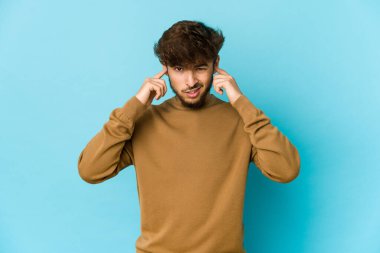 Mavi arka plandaki genç Arap adam kulaklarını parmaklarıyla kapatıyor, gürültülü bir ortam yüzünden stresli ve çaresiz..