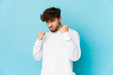 Mavi arka planda genç bir Arap, kameraya yumruk gösteriyor, agresif yüz ifadesi..