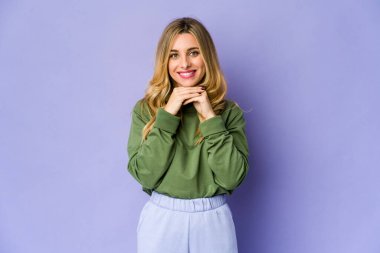 Beyaz tenli sarışın kadın ellerini çenesinin altında tutuyor ve mutlu bir şekilde yan tarafa bakıyor..