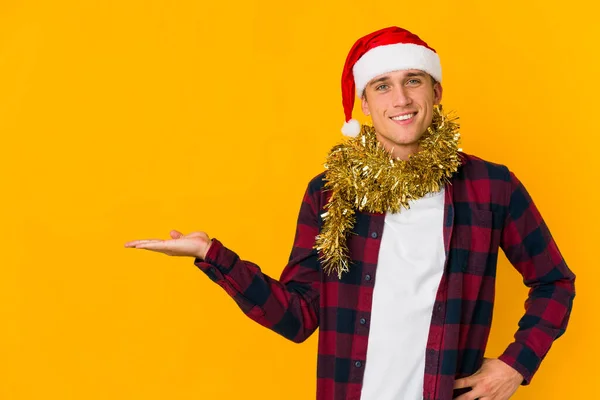 若いですCaucasian男とともにクリスマス帽子保持プレゼント隔離上黄色の背景ショーをコピースペース上の手のひらと保持別の手上の腰 — ストック写真