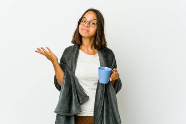 Beyaz arka planda battaniyeli genç Latin bir kadın sorgulama sırasında omuz silkiyor ve şüphe ediyor..