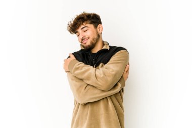 Beyaz arka plandaki genç Arap adam sarılıyor, kaygısız ve mutlu gülümsüyor..