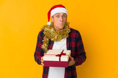 Sarı arka planda izole edilmiş bir hediye tutan Kafkasyalı genç adam omuzlarını silker ve gözleri açık kalır..