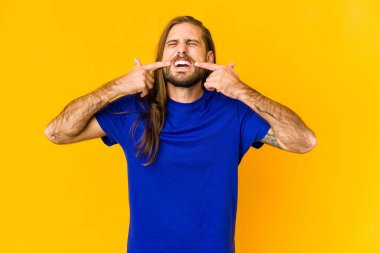 Uzun saçlı genç adam gülümsüyor, parmaklarıyla ağzını işaret ediyor..