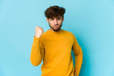 Mavi arka planda genç bir Arap, kameraya yumruk gösteriyor, agresif yüz ifadesi..