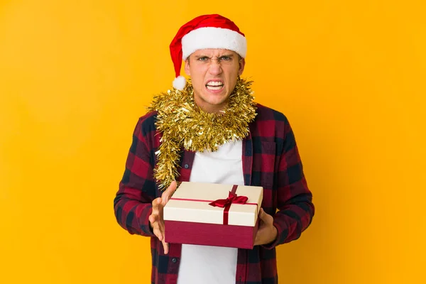 若いですCaucasian男とともにクリスマス帽子保持していますプレゼント隔離された上に黄色の背景悲鳴非常に怒って積極的 — ストック写真