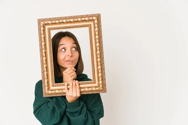 Young hispanic woman holding an old frame looking sideways with doubtful and skeptical expression.
